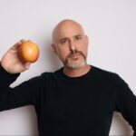 Dylan Stewart, the renowned Youtuber often dubbed "The MacWhisperer," stands against a plain white background, holding an apple in his right hand. With his bald head and distinguished goatee, he exudes a unique blend of charisma and expertise.